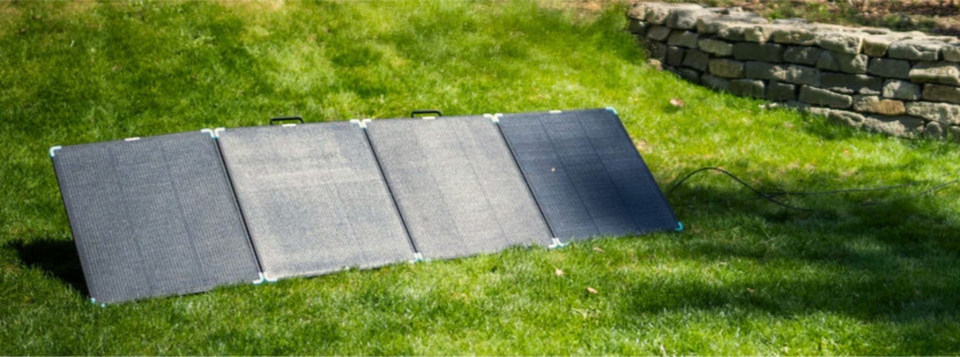 A tilted four-section folding solar panel on a lawn beside a stone wall, with dappled tree shade across the grass on the right reaching the right-hand end section, which sits darker than the three in full sun