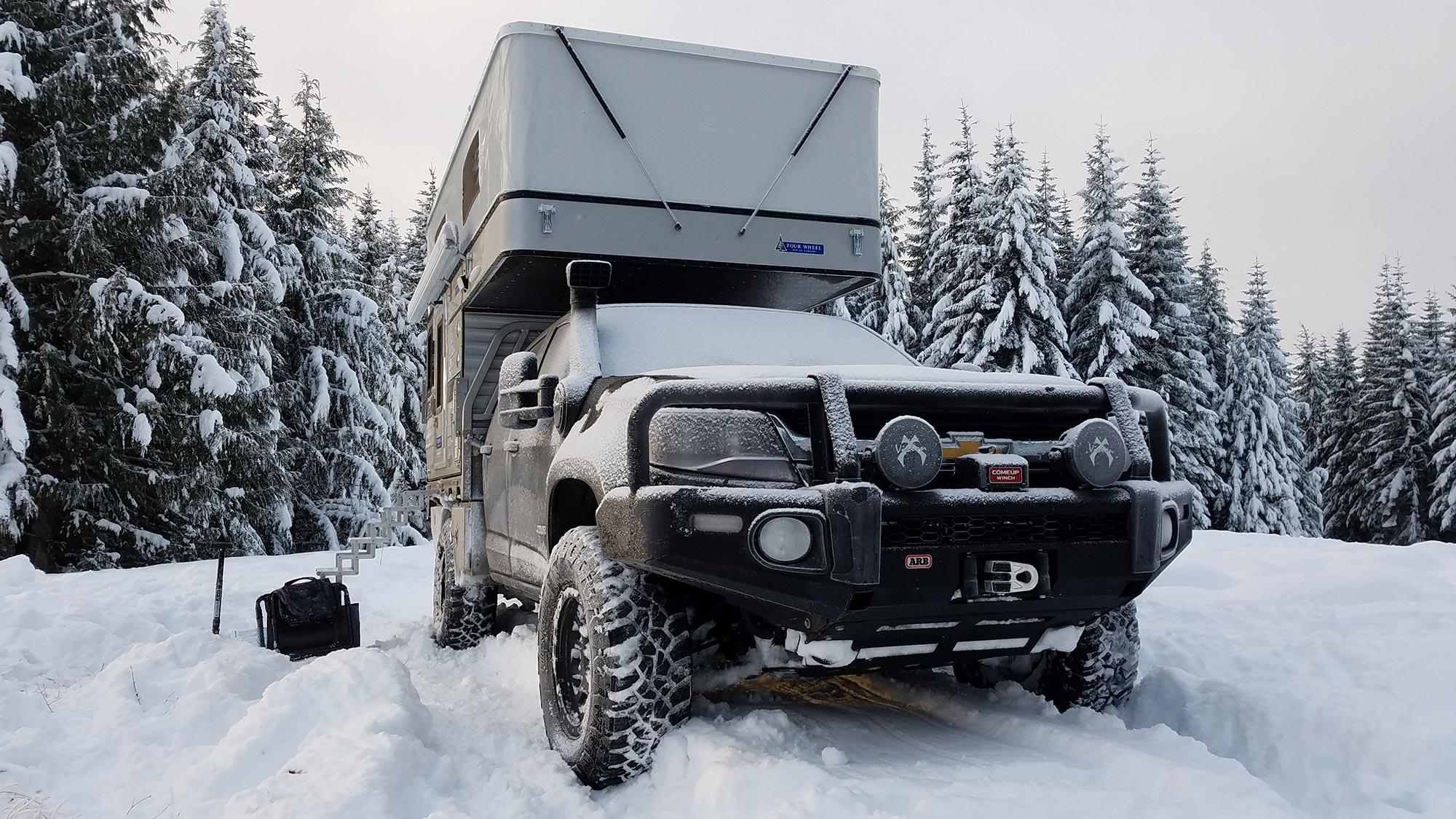 Slide-on camper on a 4WD parked in deep snow among snow-laden pines, under flat overcast winter light